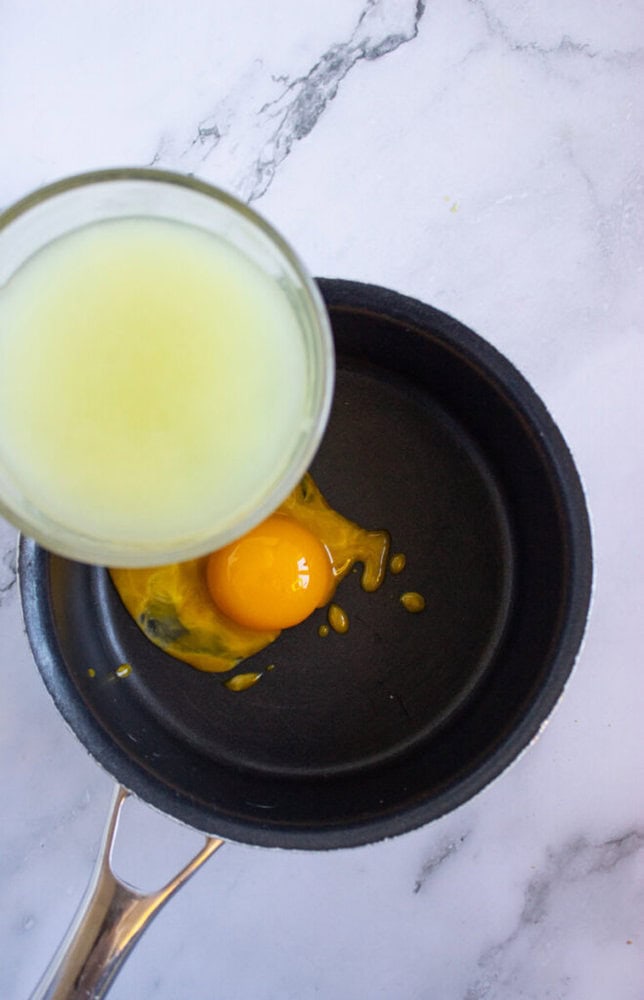 Egg and lemon juice in a pan on a marble surface, with lemon juice being poured into the pan.