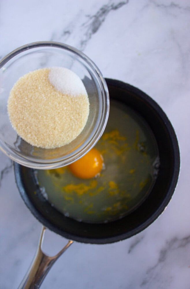 A saucepan with an egg and liquid, and a glass bowl holding granulated sugar above it.