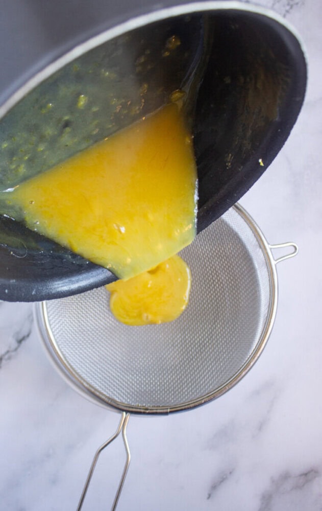 Yellow liquid being poured from a dark pan into a fine-mesh strainer over a white surface.