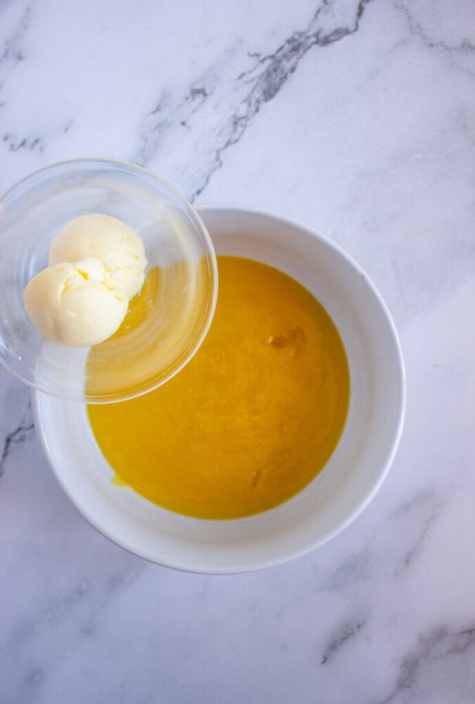 Butter being added to a bowl of melted butter on a marble countertop.