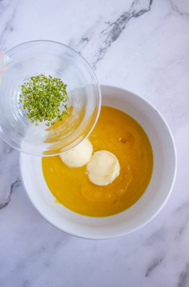A white bowl with yellow liquid and two scoops of ice cream. A hand is pouring green zest from a small glass bowl into it.