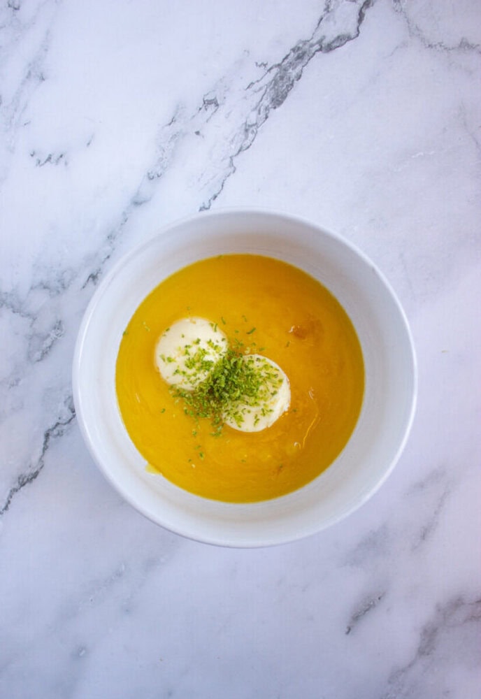 A bowl of creamy yellow soup garnished with two dollops of sour cream and sprinkled with green herbs, set on a marble surface.