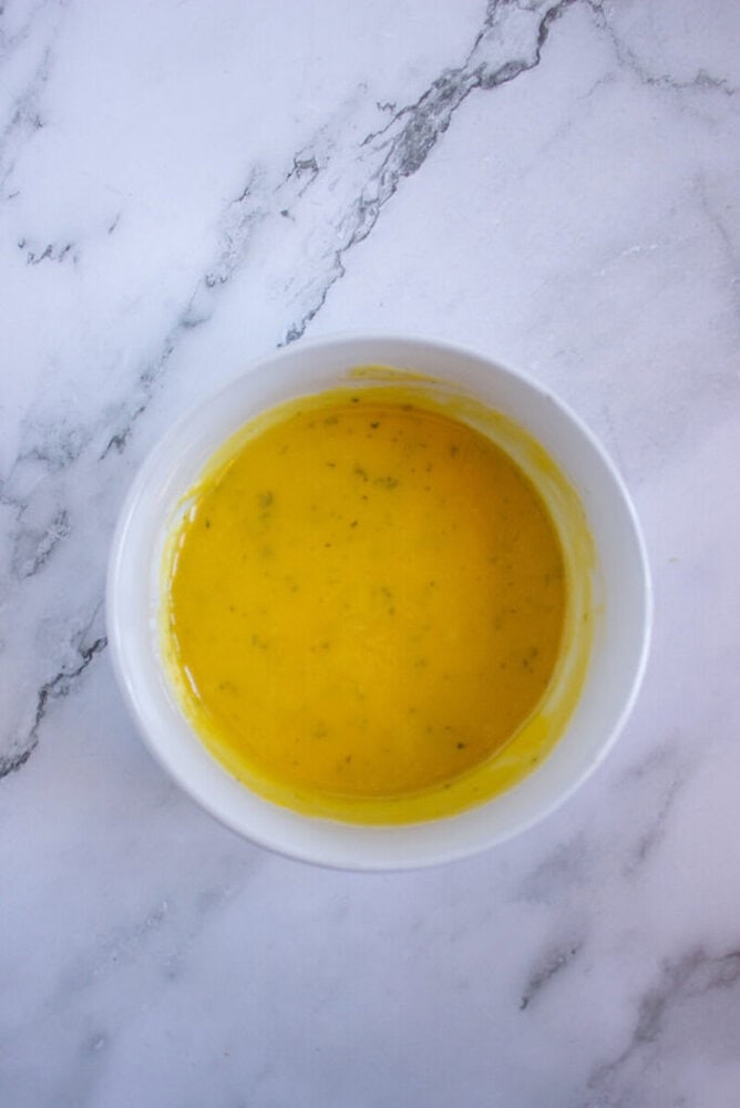 A white bowl containing a yellow liquid, likely a sauce or dressing, is placed on a marble surface.