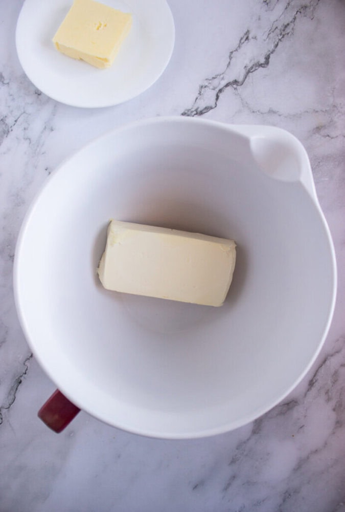 Butter in a white mixing bowl on a marble surface, with another piece of butter on a small plate in the background.