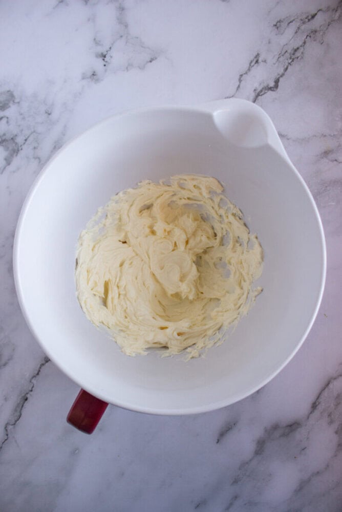 A white mixing bowl on a marble surface contains creamy, whipped butter.