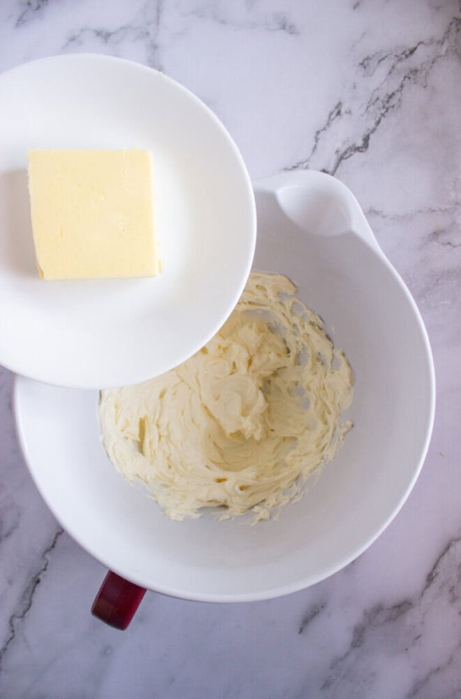 Butter block on a plate next to a mixing bowl containing whipped butter on a marble countertop.