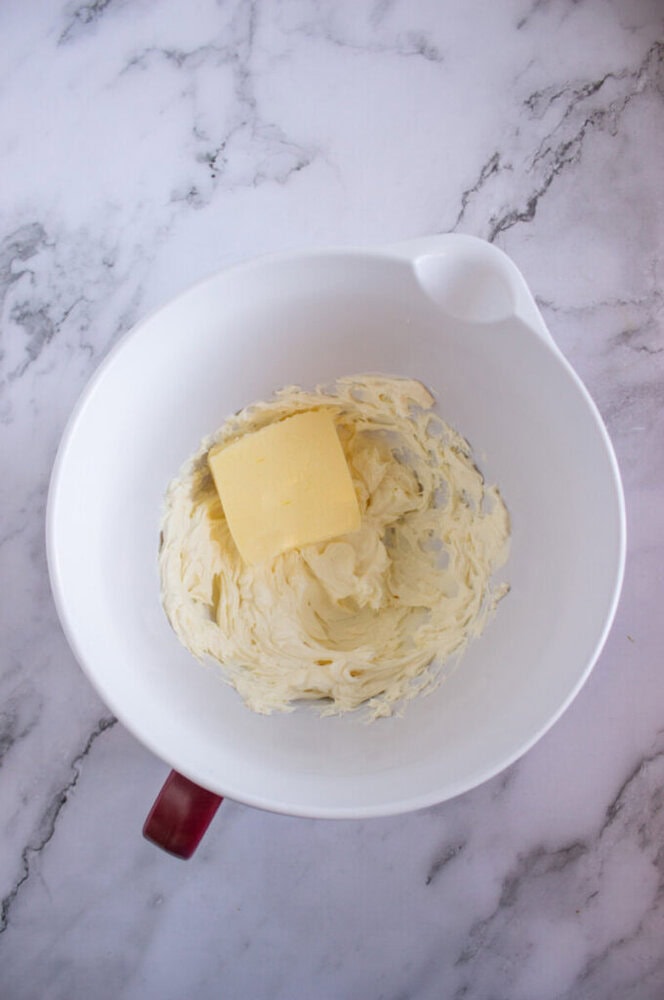 A piece of butter sits on whipped cream in a white bowl with a red handle, placed on a marble countertop.