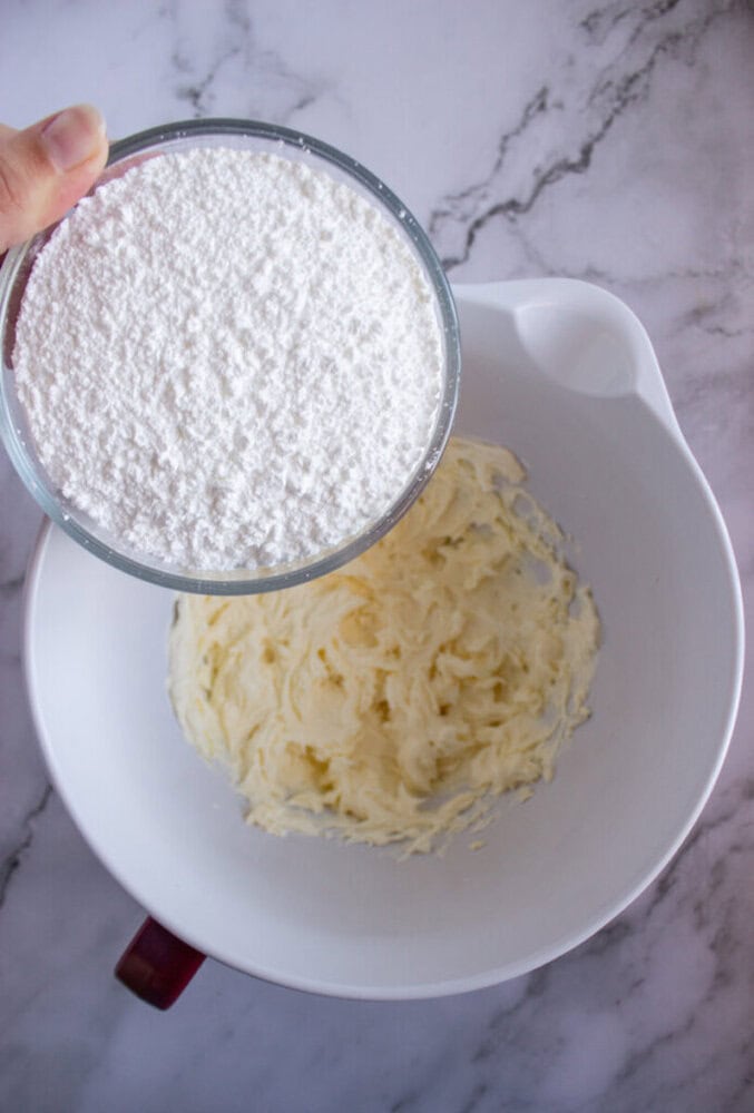 A hand holding a bowl of powdered sugar over a mixing bowl with creamed butter.