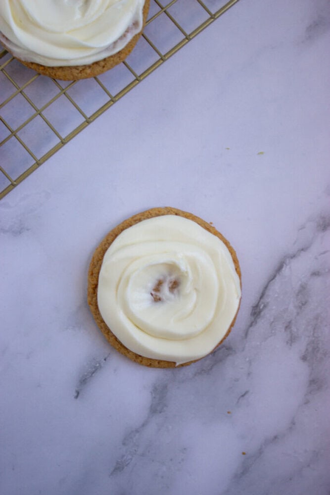 A round cookie with white frosting on top sits on a marble surface, partially placed on a metal cooling rack.