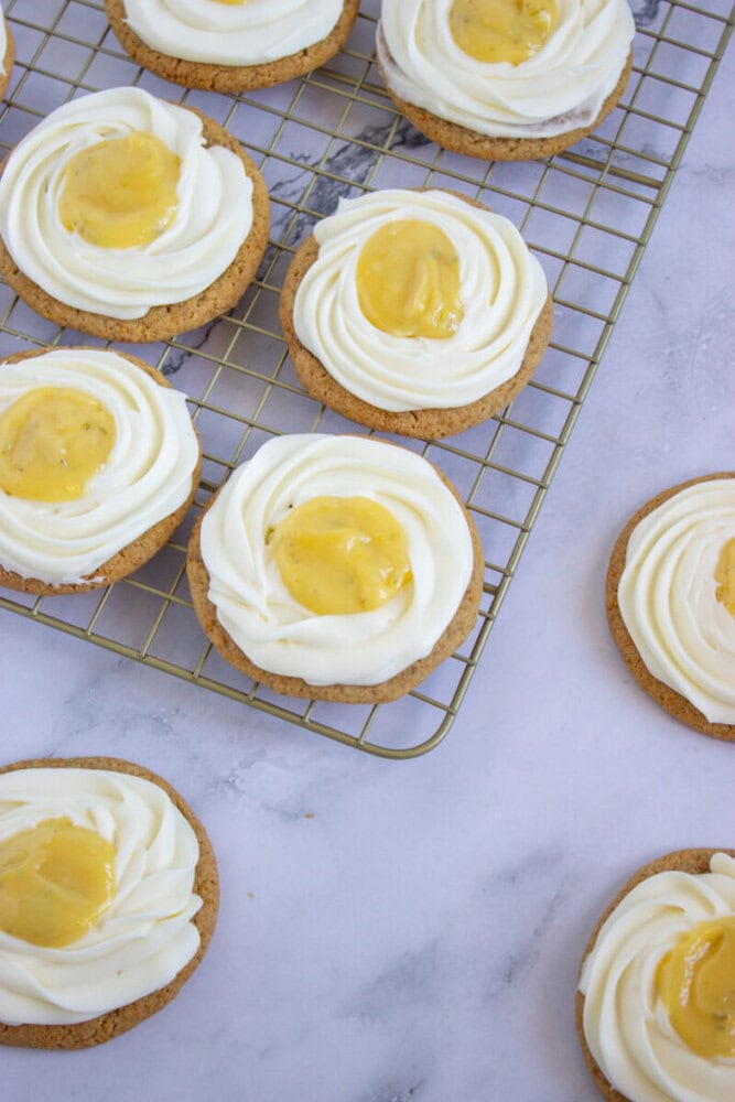 Several cookies topped with white frosting and a yellow dollop are arranged on a cooling rack and marble surface.