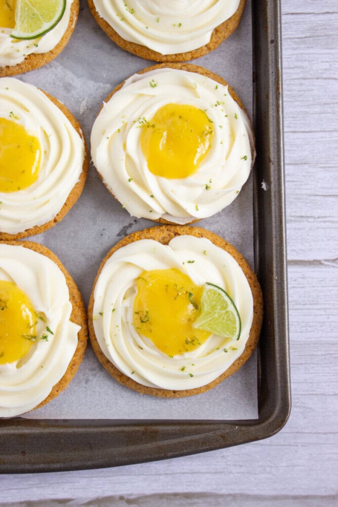 Cookies with swirled white frosting, a dollop of yellow topping, and a small lime slice, arranged on a baking tray.