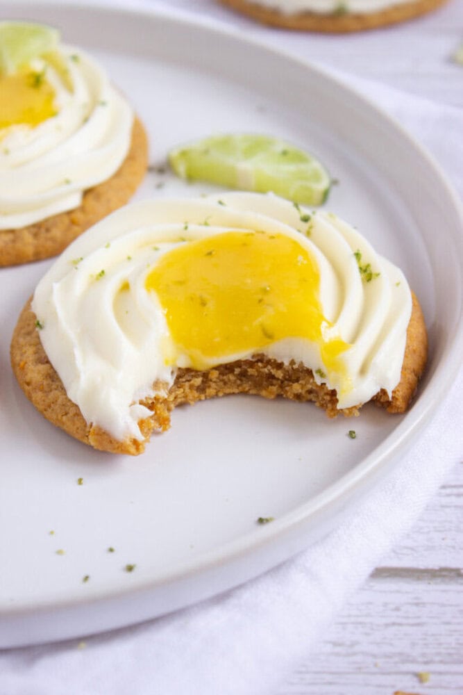 A cookie topped with white frosting and yellow lemon curd, partially eaten, sits on a plate with a lime slice garnish.