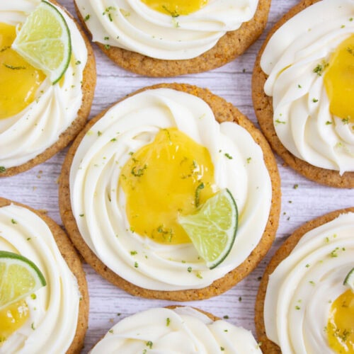 Cookies topped with swirls of white frosting, a dollop of yellow curd, and garnished with lime wedges and zest.