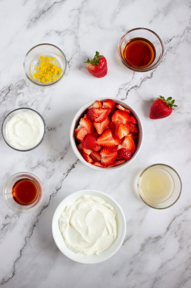 Bowls with strawberries, yogurt, lemon zest, and liquids are arranged on a marble surface with two whole strawberries beside them.