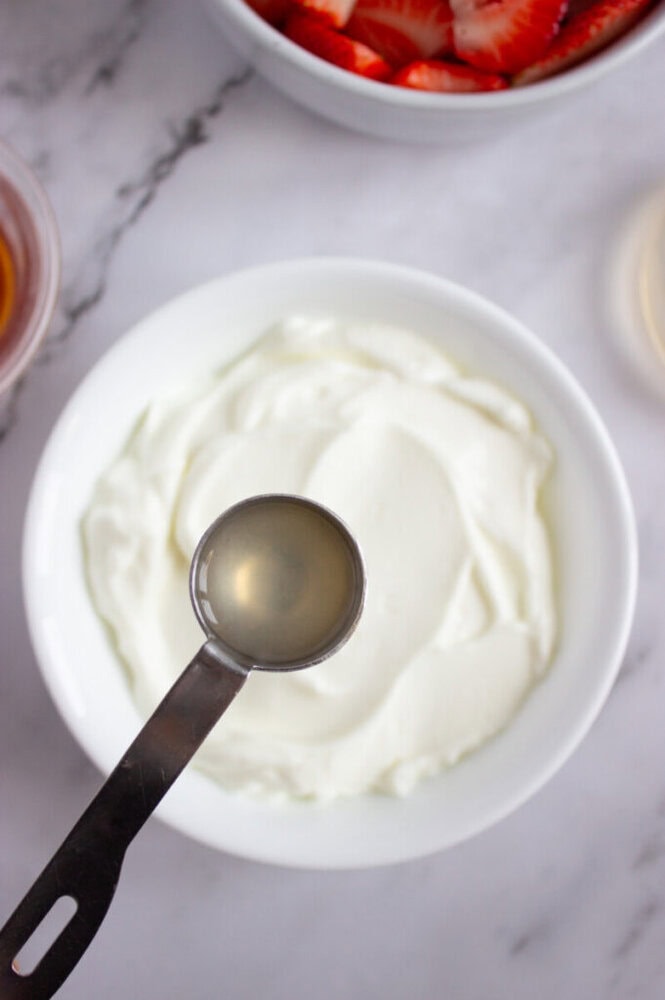 A measuring spoon with liquid held above a bowl of yogurt, with sliced strawberries in a separate bowl nearby on a marble surface.