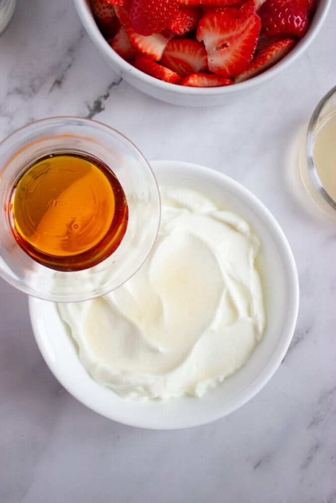 A bowl of yogurt, a glass of honey, sliced strawberries in a bowl, and a small glass of juice on a marble surface.