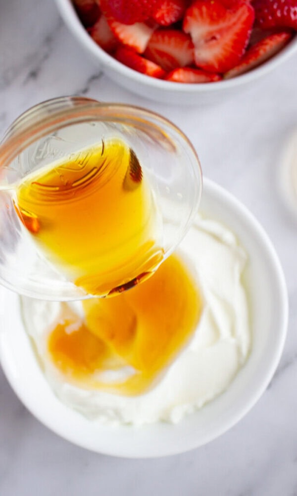 Honey being poured into a bowl of yogurt with a bowl of sliced strawberries in the background.