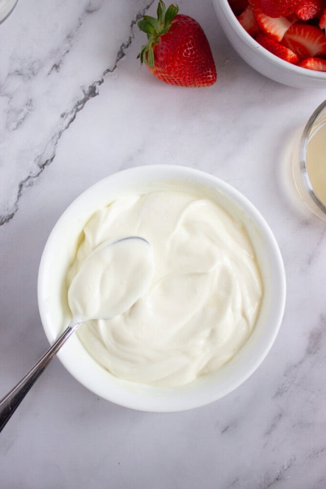 A bowl of plain yogurt with a spoon, placed on a marble surface, surrounded by whole and sliced strawberries.