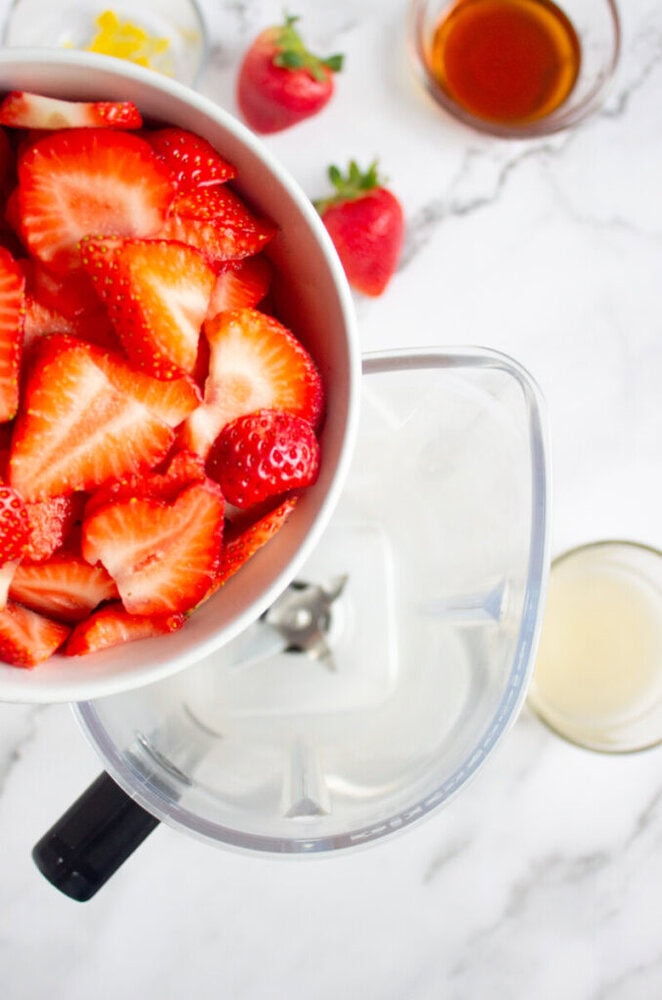 Bowl of sliced strawberries is being poured into a blender. Other ingredients and a whole strawberry are in the background on a marble surface.