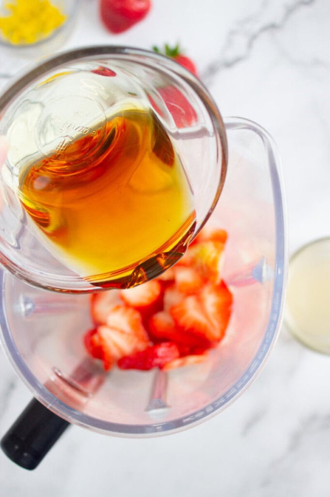 Pouring honey into a blender with sliced strawberries on a marble countertop.