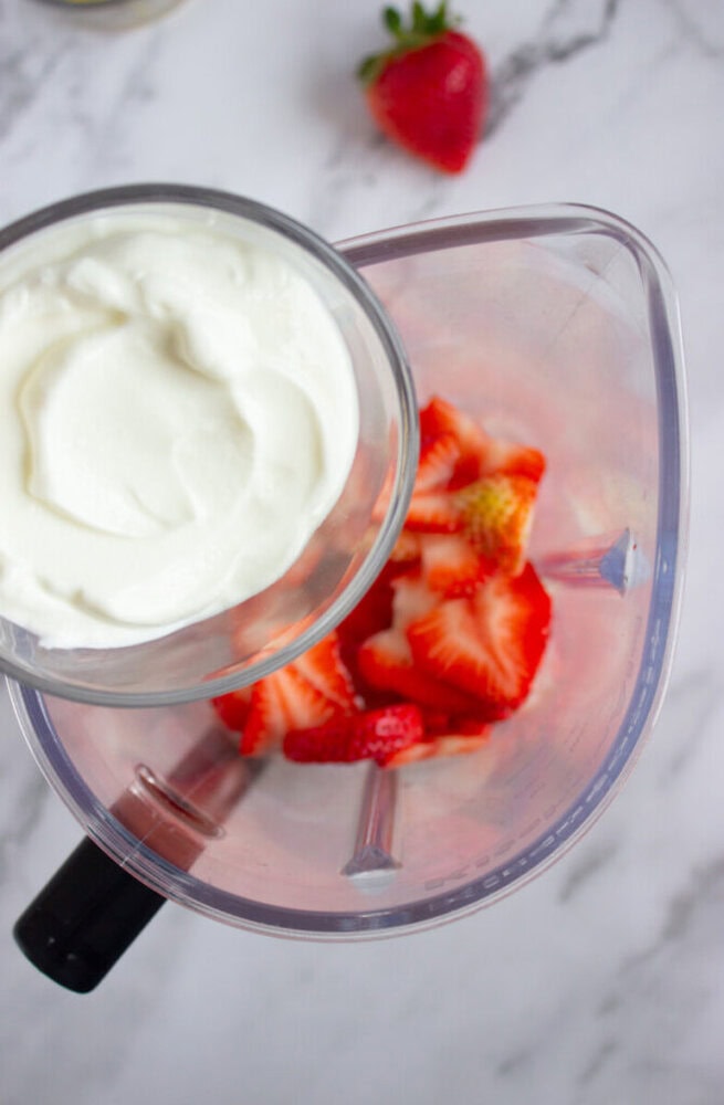 A blender with sliced strawberries and a bowl of yogurt on a marble surface. A whole strawberry is in the background.