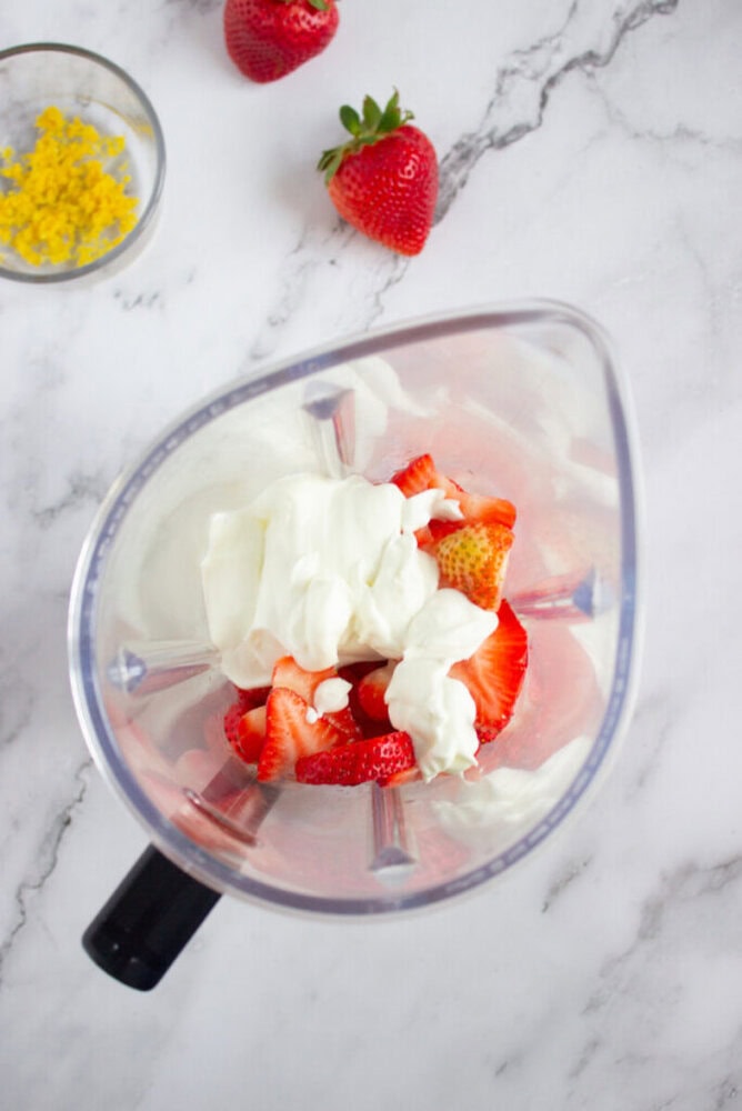 Top view of a blender containing yogurt and sliced strawberries, on a marble countertop. A small dish with lemon zest and whole strawberries are nearby.