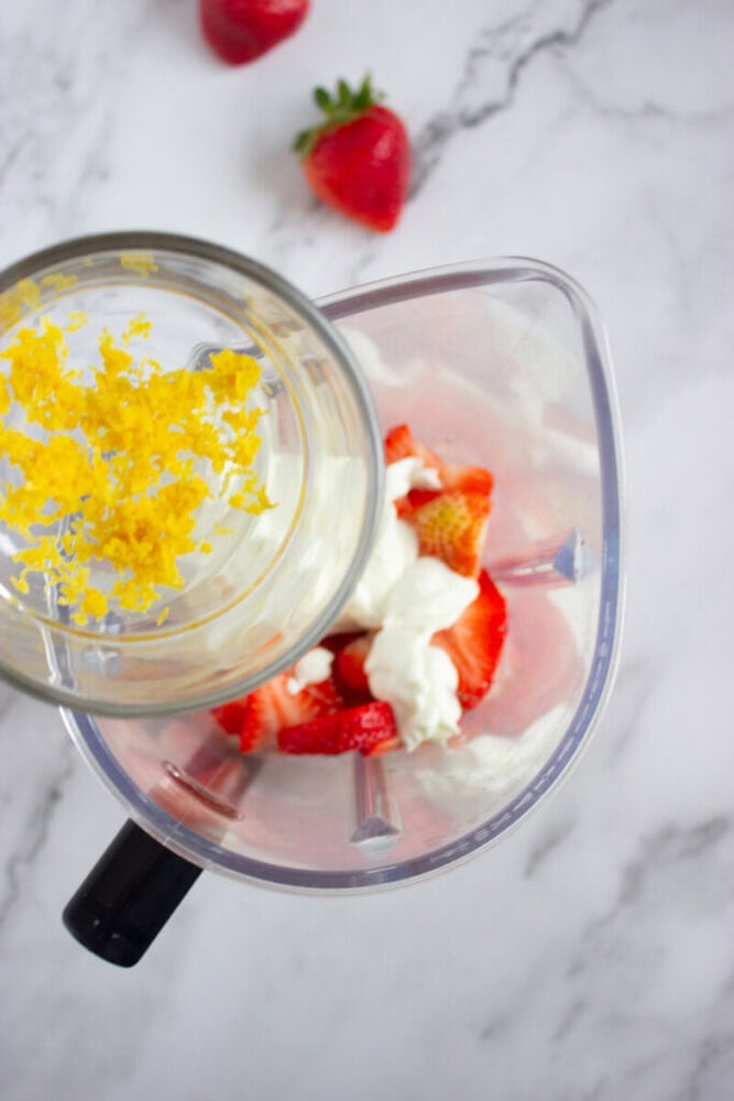 Top view of a blender with lemon zest, strawberries, and yogurt inside. Two strawberries are in the background on a white marble surface.