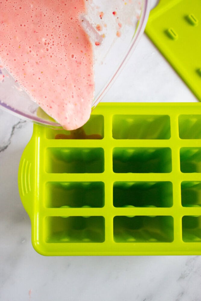 Pink liquid being poured from a clear container into a green silicone popsicle mold on a marble surface.