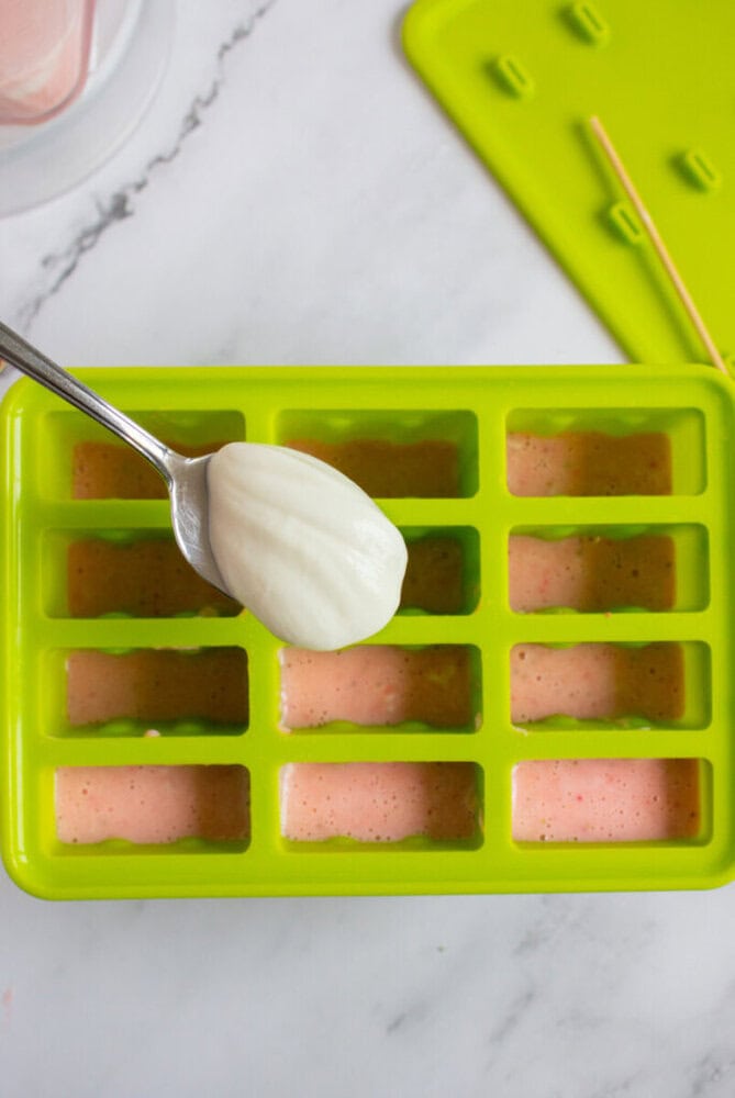 Spoon adding white mixture into green silicone mold filled with pink mixture on a marble surface, with a green lid nearby.