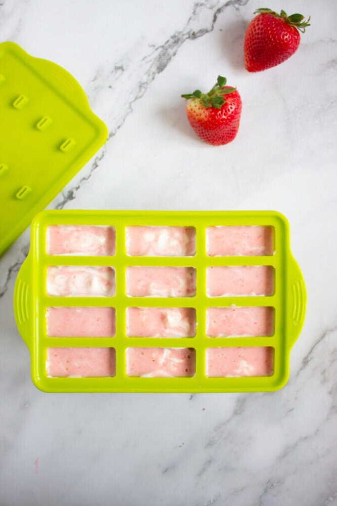 Green silicone mold filled with pink mixture on a marble surface. Two strawberries and a matching green lid are nearby.
