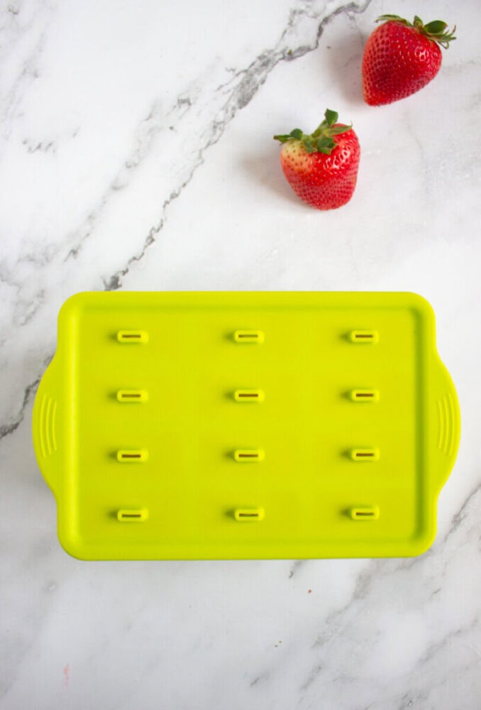 A green rectangular silicone ice cube tray with raised sticks on a marble surface. Two strawberries are placed above the tray.