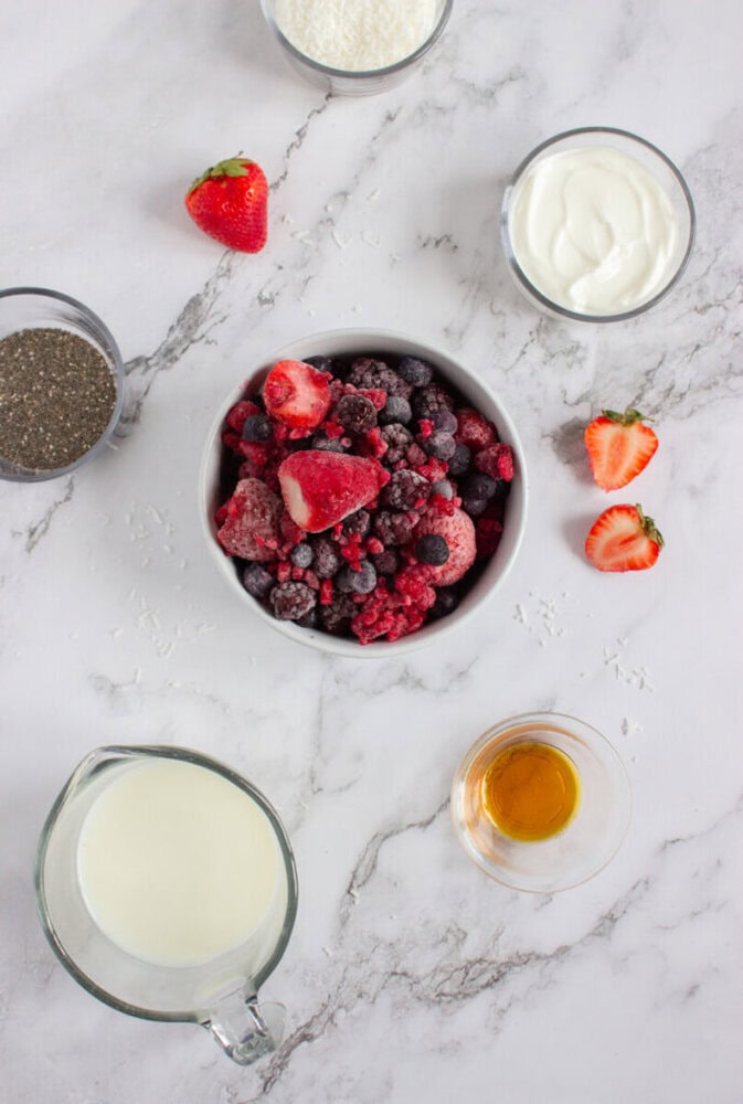 Overhead view of ingredients on a marble surface: frozen berries, chia seeds, shredded coconut, yogurt, fresh strawberries, milk in a pitcher, and a small bowl of honey or syrup.