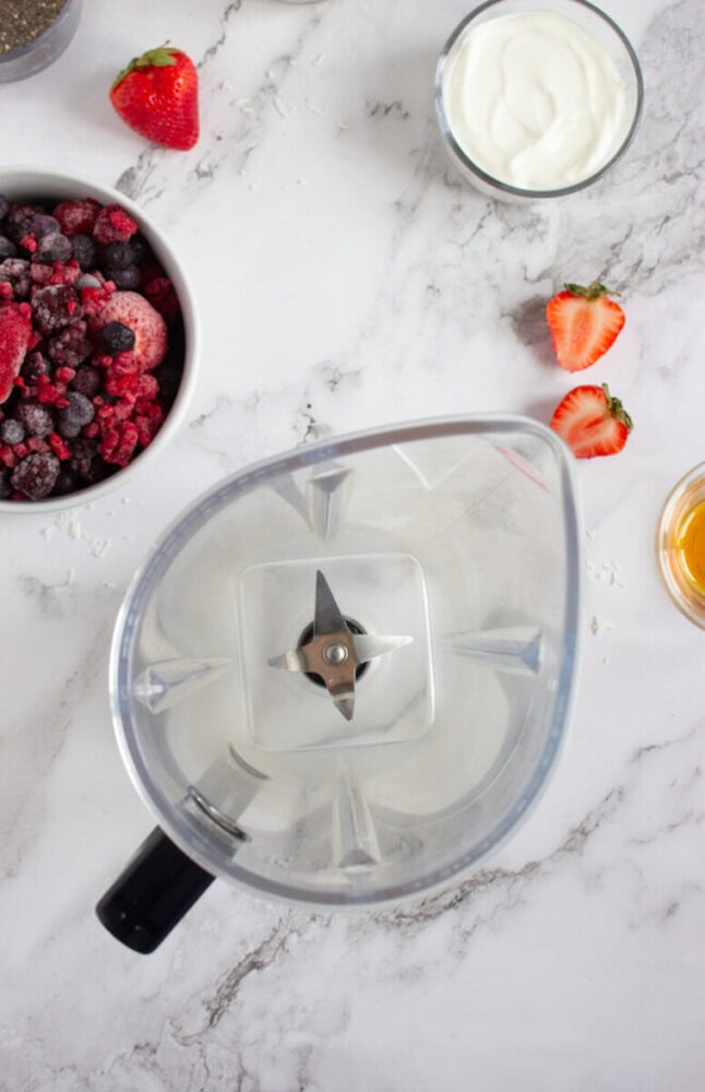 Blender jug on a marble counter surrounded by a bowl of mixed berries, sliced strawberries, a bowl of yogurt, and a small dish of honey.