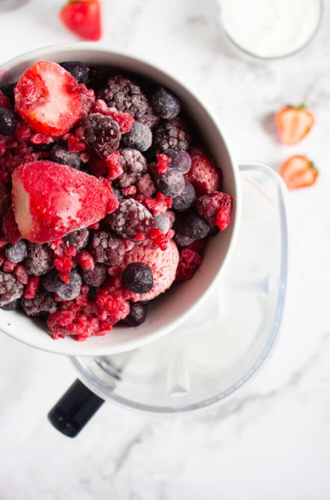 A bowl of mixed frozen berries, including strawberries, raspberries, and blueberries, sits on top of a blender. Some fresh strawberries and a glass of yogurt are in the background.