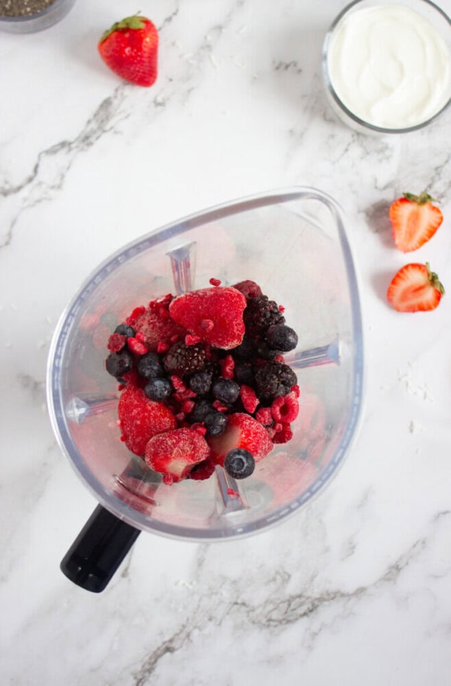 Frozen mixed berries in a blender on a marble countertop, surrounded by a fresh strawberry, halved strawberries, a bowl of yogurt, and a chia seed container.
