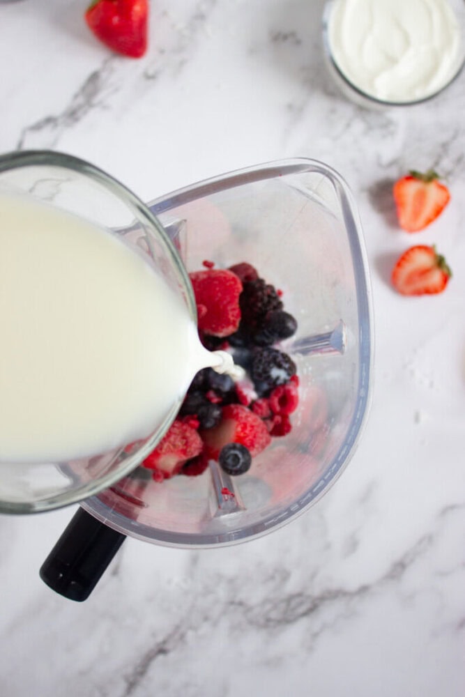 Milk being poured into a blender containing strawberries, blueberries, and blackberries. Other strawberries and a bowl of yogurt are nearby on a marble countertop.