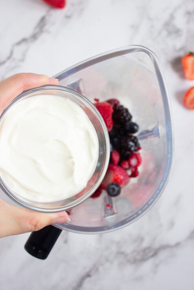 A person is holding a bowl of yogurt above a blender containing mixed berries on a marble countertop.