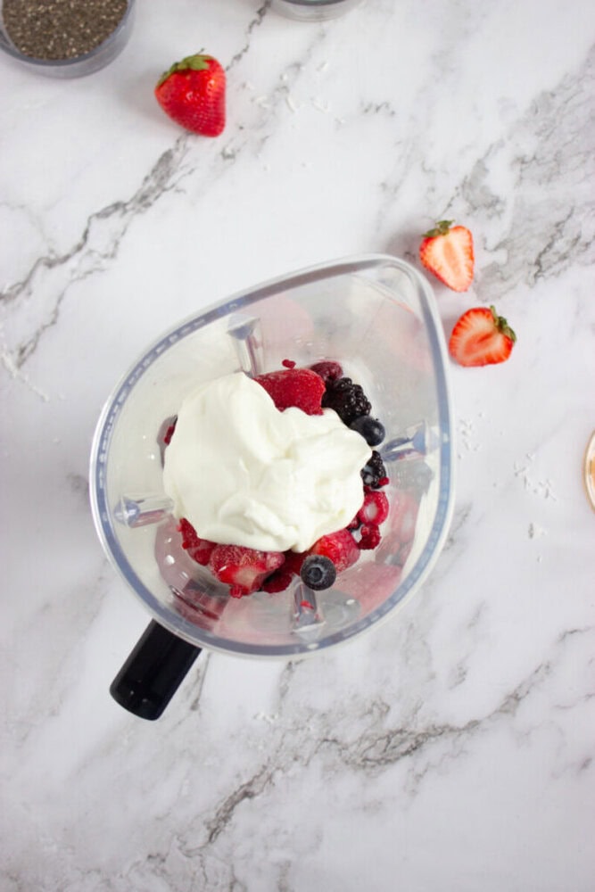 Blender with assorted berries and a dollop of yogurt, on a marble surface with a whole and sliced strawberry nearby.