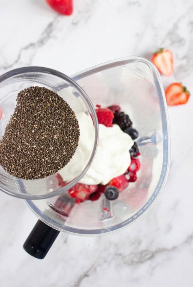 Blender with yogurt and mixed berries, a hand adding chia seeds from a bowl, on a marble countertop.