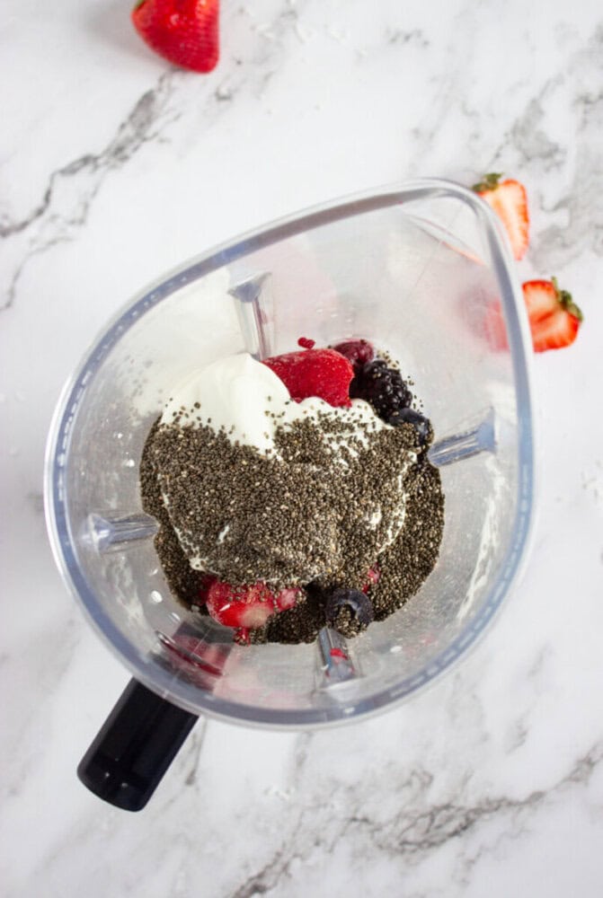 Blender with chia seeds, yogurt, and mixed berries inside on a marble countertop.