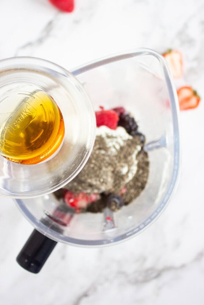 Pouring liquid into blender with chia seeds, berries, and other ingredients on a marble countertop.