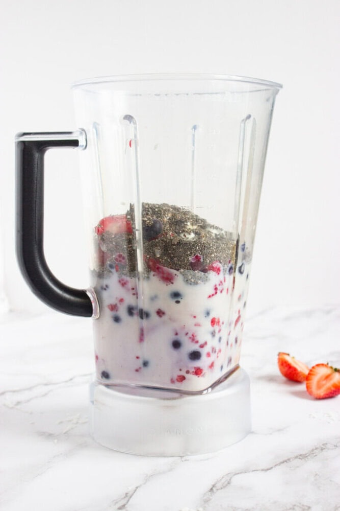 Clear blender contains milk, berries, and chia seeds, placed on a white marble surface. A few sliced strawberries are next to it.