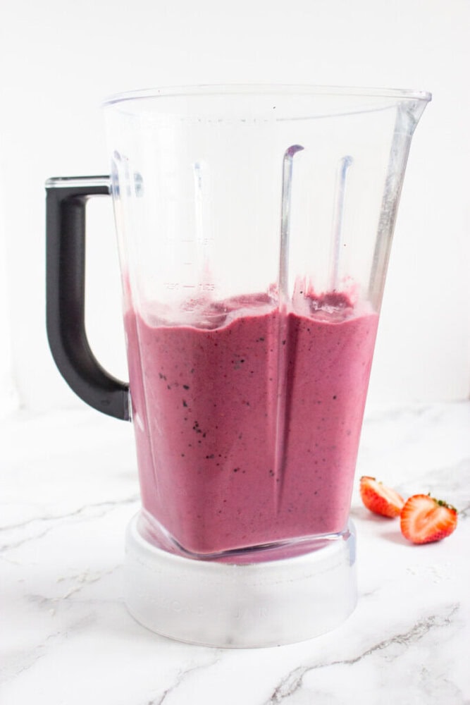 A blender filled with a thick, pink smoothie sits on a marble countertop. Two halved strawberries are placed beside the blender.