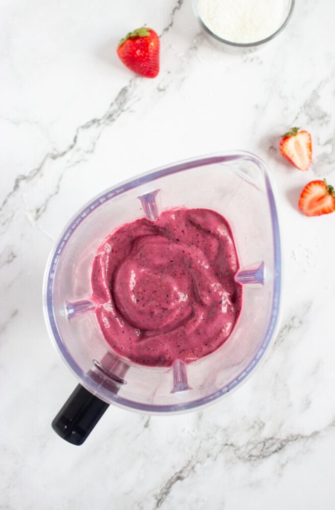 A blender filled with thick, purple smoothie sits on a marble surface. Nearby are whole and halved strawberries and a bowl of grated coconut.