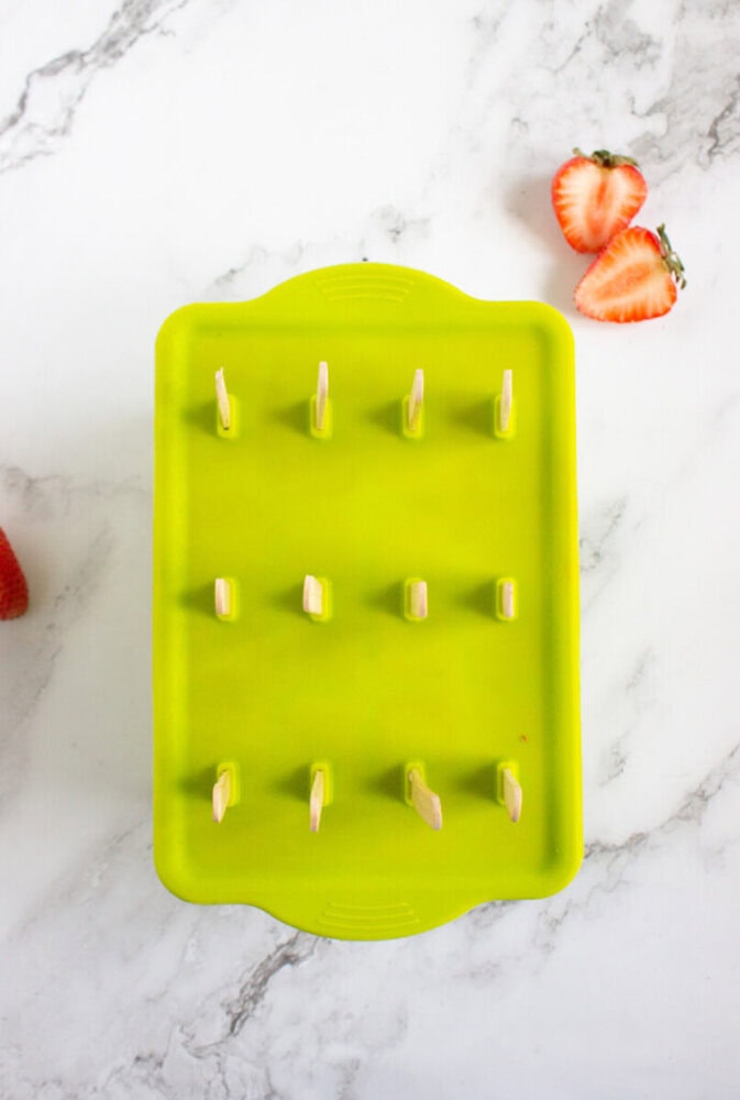 Green popsicle mold with wooden sticks protruding, placed on a marble surface with halved strawberries nearby.