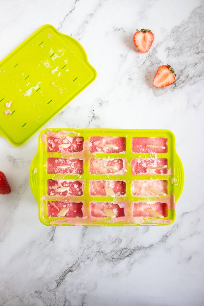 Ice cube tray filled with pink, partially frozen liquid on a marble surface, with a lid and halved strawberries nearby.