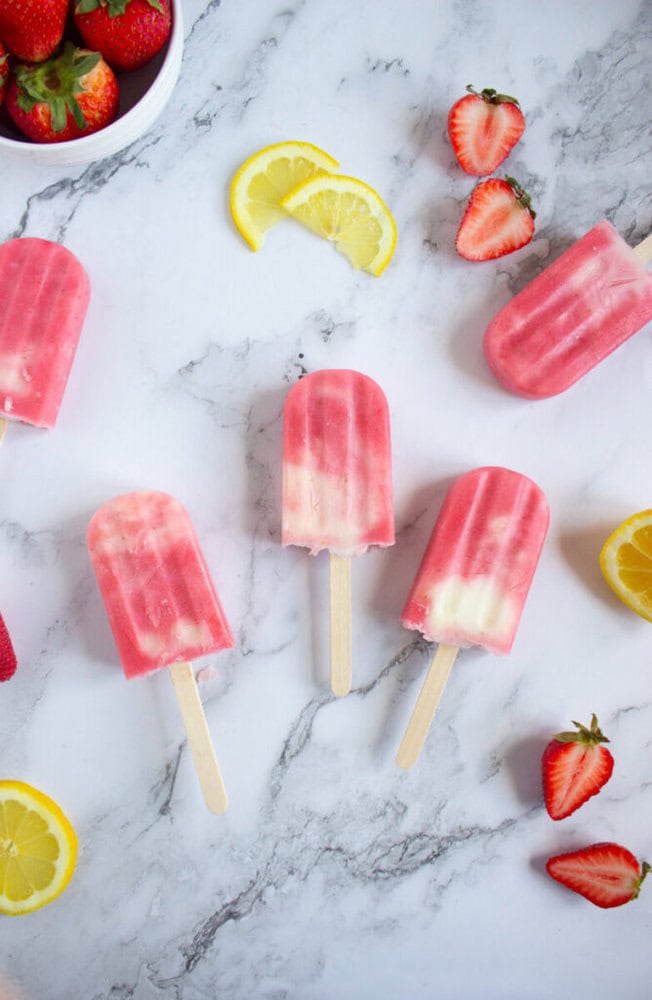 Five pink and cream popsicles arranged on a marble surface with lemon slices and fresh strawberries scattered around.