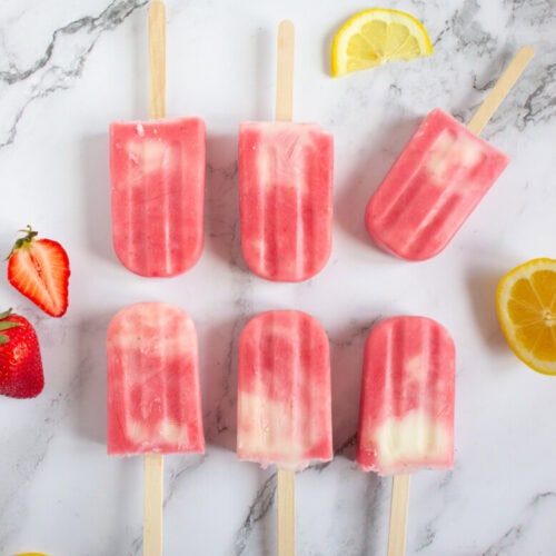 Six pink popsicles with visible swirls on wooden sticks are arranged on a marble surface, surrounded by sliced strawberries and lemon wedges.