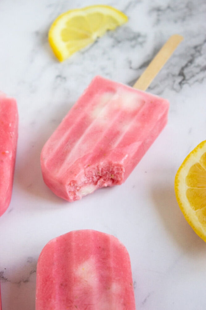 Pink popsicles with a bite taken out of one are on a marble surface, surrounded by lemon slices.