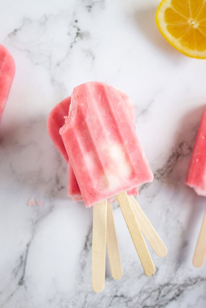 Two pink and white popsicles with a bite taken from one, on a marble surface, next to a lemon slice.